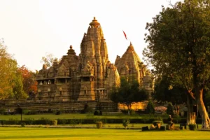 Intricately carved stone architecture of a temple within the Khajuraho Group of Monuments, surrounded by a green lawn and trees at sunset.