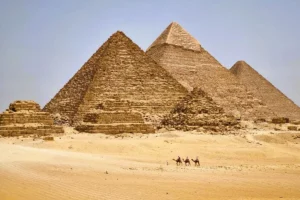 A view of camel riders crossing the sandy Giza Plateau towards the three Great Pyramids of Giza (Khufu, Khafre, and Menkaure) near Cairo, Egypt.