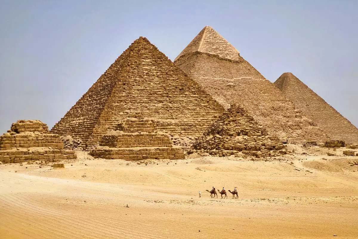 A view of camel riders crossing the sandy Giza Plateau towards the three Great Pyramids of Giza (Khufu, Khafre, and Menkaure) near Cairo, Egypt.