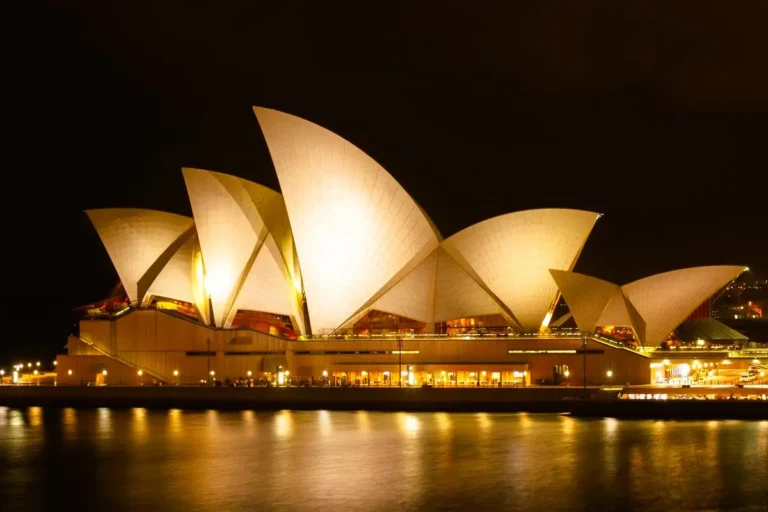 The illuminated shell-shaped architecture of the Sydney Opera House glowing at night by the harbor water